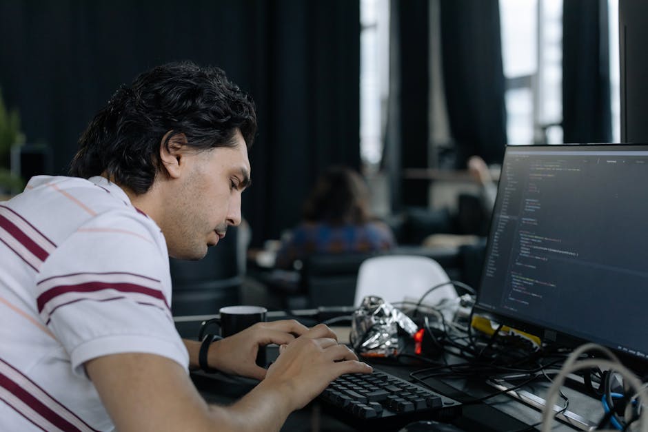 A young man with dark, wavy hair wearing a white t-shirt with red and beige horizontal stripes is working on a computer in an indoor office setting. He is focused on the screen, which displays lines o